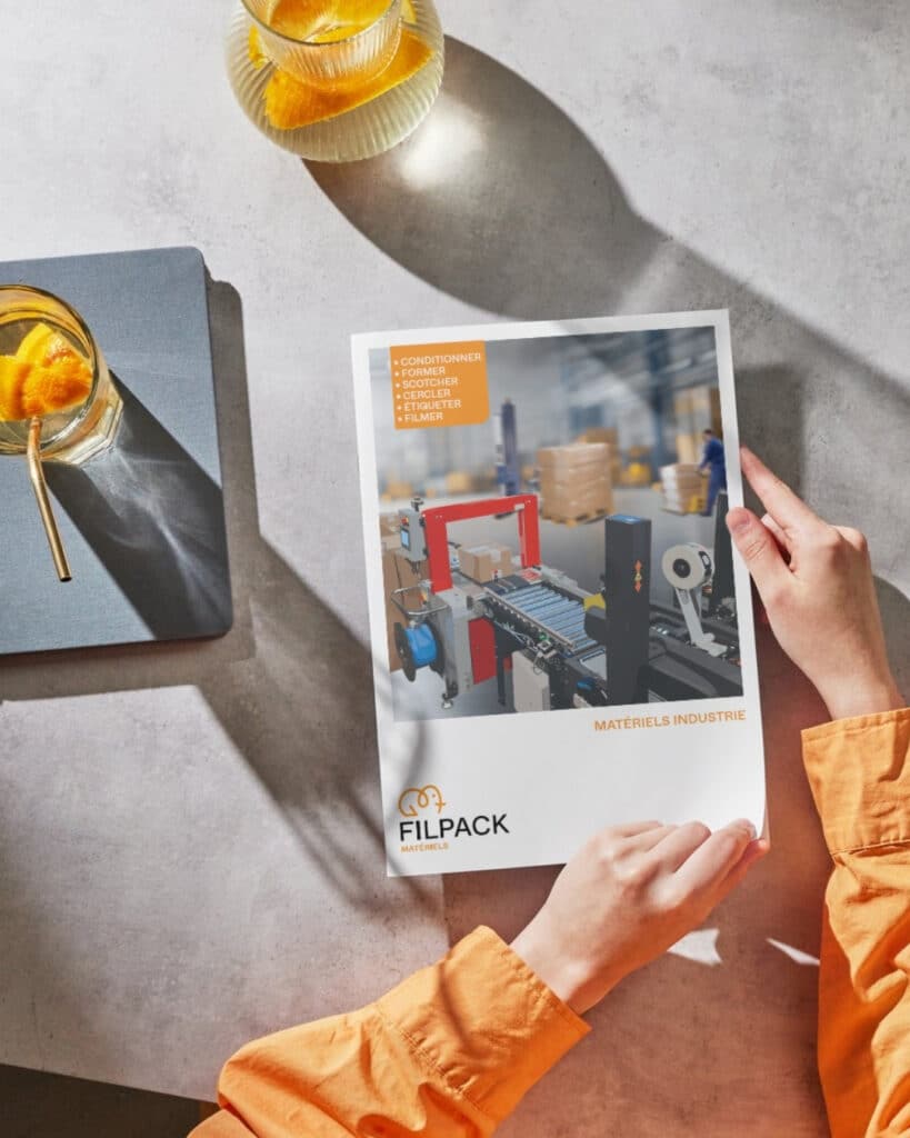 Catalogue Filpack Matériels Industrie 2025 tenu par une personne, présentant des machines industrielles pour le conditionnement et l’emballage dans un entrepôt logistique.